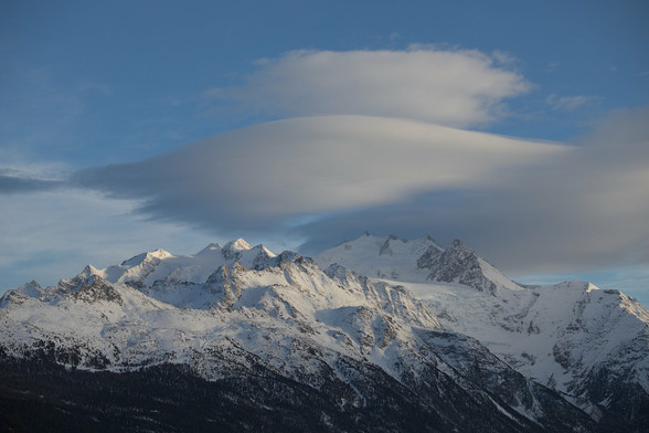 Berglandschaft im Wallis: Blauer Himmel mit Föhnfischen über den Berggipfeln des Mischabel-Massivs. Die Berge sind noch eingeschneit, die Baumgrenze unterhalb ist dunkel-braun.
---
Mountain landscape in Valais: Blue skies with cumulus lenticularis clouds over the peaks of the Mischabel massif. The mountains are still covered in snow, the tree line below is dark brown.