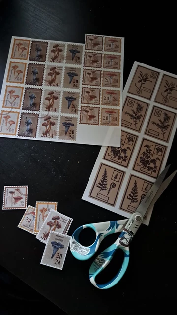 Two printed sheets of stickers being cut by hand. The scissors are on the black table top next to the sticker sheets. The stickers on the first sheet are stamp shaped with mushroom illustrations on them. Secon smaller sheet has botanical illustrations.