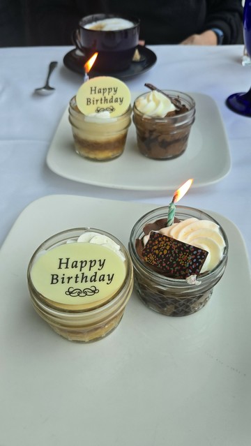 Little cups of dessert,  one is white chocolate and days Happy Birthday. The other is dark chocolate with a blue white candle and a fan of whipped

On white, square plates on a white table cloth