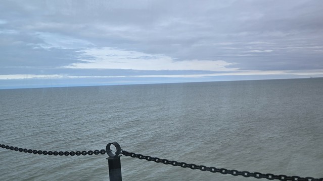 View of grey green lake Erie out the windiest with a chain fence in the foreground. The sky is overcast with some spots of blue