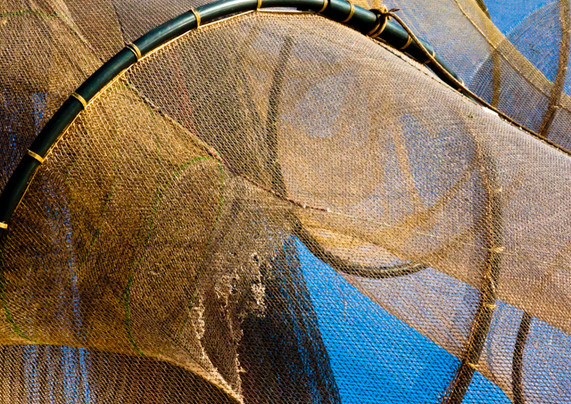 Nets on a large, historic sailing ship. There is a large black ring, you can see three quarters of it. The ring runs from the left, along the top of the photo to the bottom right. A brown net is attached to the ring all around. In some places you can look through it and see the blue sky. Behind it are more of these nets stretched. A net on the left is frayed. It looks like jute, but very thin. At the top right you can see a few more of those black rings with nets on them.