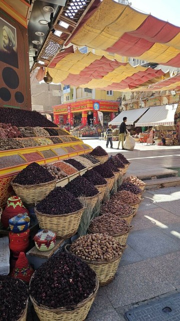 Photo showing an outside stall of a bazar. Big baskets filled with heaps of different coloured spices line the ground along the table in three different heights. Colourful little vessels with lids are placed in between. On the table are rows and rows of open little boxes with various spices and several with fresh or dried dates. Yellows, reds, orange and browns of finely grained and powdered spices reach up the wall. Above the stall are bright red and yellow cloths spanned across the tiled path to protect from the sun. In the background along the path are a lot more stalls, hidden behind their own sunshades. Bright red coloured walls are visible at the end of the alley.