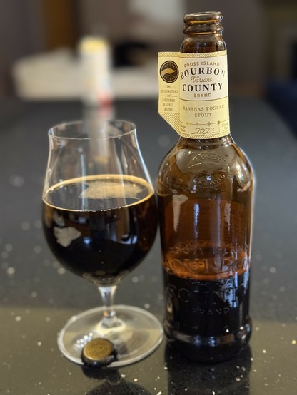A snifter glass of dark beer next to a pint brown bottle with a neck label in light yellow.