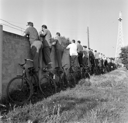 A monochrome photo of about 20 men standing on their bicycles that are leaning against a high concrete brick wall. They are peering over the fence and although we can't see what they are looking at, the provenance of the photo tells us they are watching a football match on the other side of the wall. Some of the men are in short-sleeved shirts, suggesting it is a fine day.