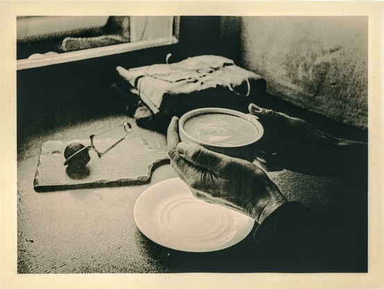This is a black-and-white lith print photograph, a darkroom printing technique known for its high contrast, deep blacks, and warm tones. The image depicts a close-up of persons hands holding a cup of coffee with intricate latte art on its surface. The hands are detailed and textured, emphasizing the tactile quality of the moment. 

In the background, a table setting includes a white plate, a textured wooden surface, and what appears to be a tea strainer and a cutting board. A folded cloth rests near the edge of the table. The lighting is soft and diffused, creating an intimate, cozy atmosphere. The grainy texture and high contrast of the lith print technique enhance the nostalgic and artistic feel of the image.