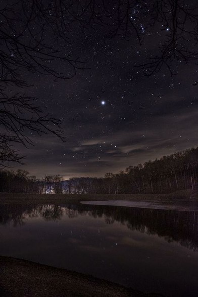 Nearly dark image, with a starry sky. Reflections in the water of a lake of some trees in the background. Some distant light in the horizon lights up a mountain ridge barely visible.