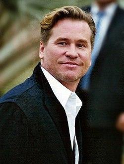 Primer plano de un hombre de mediana edad, Val Kilmer, con el pelo rubio peinado hacia atrás. Tiene una expresión seria pero amable. Viste una camisa blanca abotonada debajo de una chaqueta negra. El fondo está desenfocado, pero se puede ver a otra persona con traje oscuro detrás de él.
