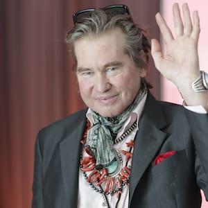 Retrato de un hombre de mediana edad, Val Kilmer, con el pelo y los ojos claros, que lleva gafas de sol sobre la cabeza. Luce un traje oscuro, una pañoleta de color verde claro alrededor del cuello y un collar con cuentas de coral rojo y turquesa. Está saludando con la mano derecha y tiene una expresión amable en la cara. El fondo es borroso y de color marrón rojizo.