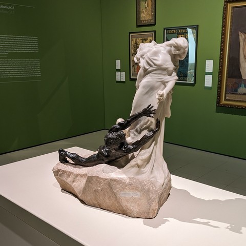Statue of a female (carved marble) with a man (cast bronze) laying at her feet reaching for the flower she holds.