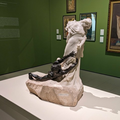Statue of a female (carved marble) with a man (cast bronze) laying at her feet reaching for the flower she holds.