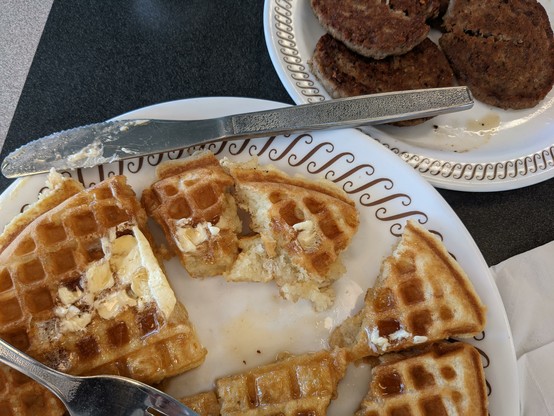 close-up of a waffle that has been cut into sections, with melted butter and syrup on top. There is another plate with several sausage patties. Waffle House