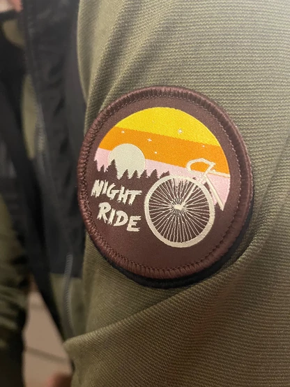 Ein Patch auf dem steht "Night Ride" mit dem stilisierten Vorderrad eines Rades mit Rennlenker vor einem Wald. Angebracht auf dem Ärmel eines grünen Trikots.