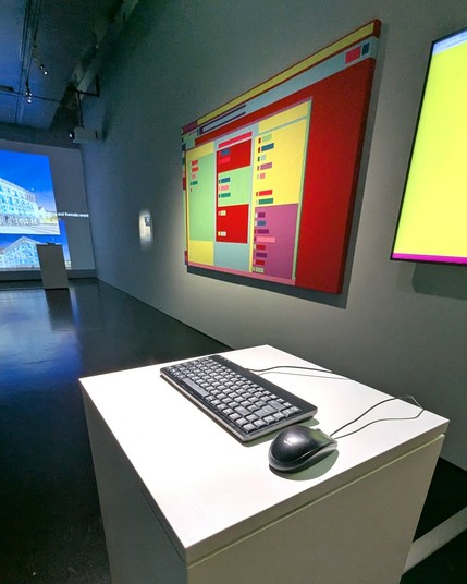 A dimly lit exhibition space with a white pedestal holding a black computer keyboard and mouse. In the background, a large, colorful artwork resembling an abstract web page layout is mounted on the wall. The colors are bold and geometric, with red, yellow, green, and blue blocks. Other screens and installations are visible in the distance.