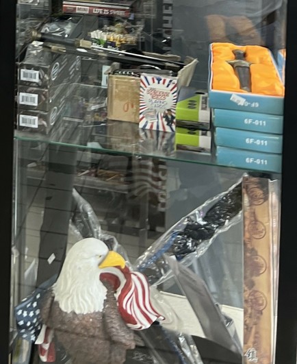 Close up of a different shelf. It has an assortment of large knives, some folding, some not. At the bottom is a plastic resin statue of a bald eagle.