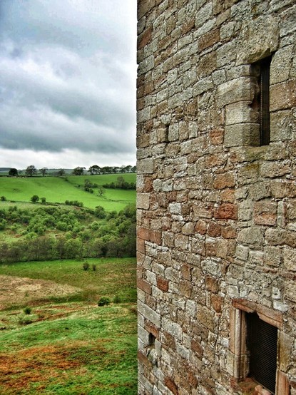 A close-up, slightly high-angle shot shows a weathered stone wall with two small, barred windows on the right side of the frame. To the left, a vast, undulating green landscape stretches out under a cloudy sky. The stone wall is constructed from irregularly shaped, light brown and grey stones, showing signs of age and weathering. The windows are small, rectangular openings with dark metal bars.
The landscape on the left is a patchwork of vibrant green fields and darker green patches of trees and bushes, suggesting rolling hills and valleys. A line of trees is visible on the horizon under a heavy, overcast sky filled with grey clouds. The overall lighting suggests a daytime scene under cloudy conditions.