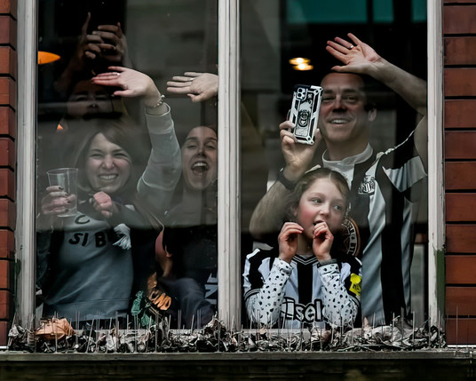 Famliy look out of a 1st floor restaurant window laughing and cheering at the football team on the open top bus. The father is in a black and white newcastle football top holding a mobile phoe to photograph the eam on teh bus. His daughter is in front of him looking pensive. Mum & Aunt are in the other window.