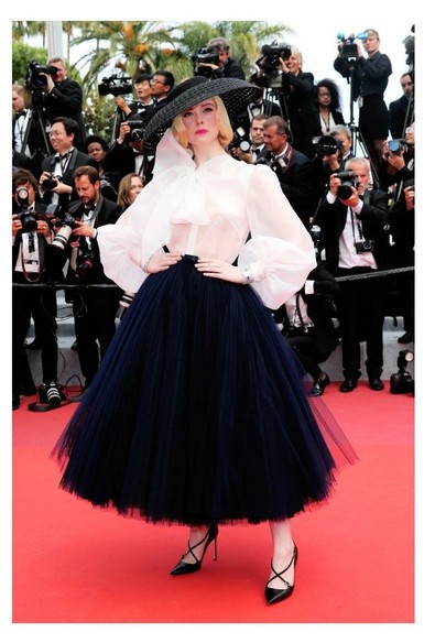 Actress Elle Fanning dressed in a beautiful outfit with oversized black hat black skirt, high heels, and frilly white top—looking fabulous, old-fashioned, and feminine for the photographers