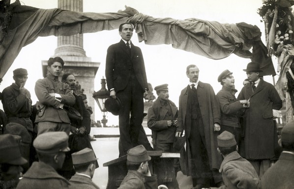 A man in a suit, holding his hat in his right hand, is addressing a crowd. He is standing on a small plinth, and the angle of the photo is looking up at him.  The few members of the crowd shown in the foreground of the photo look like soldiers. The speaker has other men standing either side of him, some of whom are dressed in early Soviet military attire. In the background is a large plinth, suggesting the place of the photo is Heroes Square.