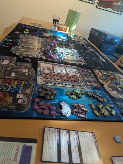 Shot from one end of the table across. Game mat with minis, board pieces, tokens, a die or two, cards, player boards. Glass and Toffifees in the background.