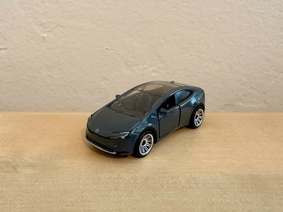 This is a small die-cast toy car model on a wooden surface, a gray Toyota Prius Prime with a hatchback design. The car has black windows and silver wheel rims. The model sits on a light brown wooden table with a beige wall visible in the background.