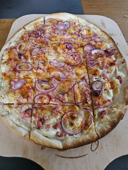 Das Bild zeigt einen Flammkuchen auf einem Holzbrett, der in acht Stücke geschnitten ist. Der Flammkuchen hat eine dünne, goldbraune Kruste und ist mit einer weißen Sauce bedeckt, die wahrscheinlich eine Art von Schmelzkäse oder cremiger Sauce ist. Der Flammkuchen ist mit roten Zwiebelringen und kleinen Würfeln von Speck garniert. Die Zwiebelringe sind in verschiedenen Größen und liegen über den Flammkuchen verteilt, während die Speckwürfel gleichmäßig verteilt sind. Die Kruste hat einige gebrannte Flecken, was auf eine hohe Backtemperatur hinweist. Der Flammkuchen ist auf einem Holzbrett platziert, die auf einem Holztisch liegt, und im Hintergrund ist ein Teil eines dunkelgrauen Tisches zu sehen.

Bereitgestellt von @altbot, privat und lokal generiert mit Ovis2-8B