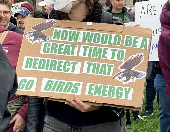 A large cardboard sign with a couple of screaming eagles in its corners, reads, in green print: "NOW WOULD BE A GOOD TIME TO REDIRECT THAT *GO BIRDS* ENERGY!"