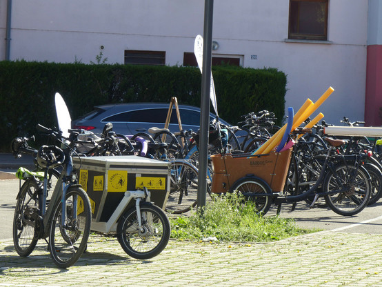 Des arceaux à vélos envahie de vélos de toute sorte. En premier plan, un vélo cargo avec des logos jaune du cadre, en arrière plan, sur les arceaux un vélo cargo avec une petite dizaine de frite de piscine qui dépassent.