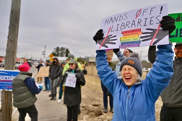 A woman holds up a sign at a rally. The sign reads, "Hands Off Libraries!" with a rainbow-colored stack of books. To the left behind her, a "Bernie Sanders for President" sign is on a telephone pole.