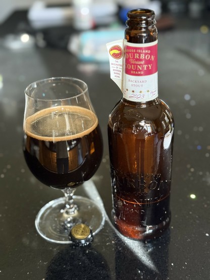 A snifter glass of dark beer next to a tall brown bottle with a white and red neck label. On the small label flag is the head of a goose.