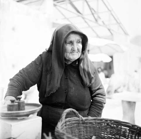 A black and white half-length portrait photo of an elderly woman wearing a largish, rough-hewn head scarf and dark clothing. She has a pensive, almost sad look on her wrinkled face. In the foreground there is a cane basket and some metal weights for weighing produce.