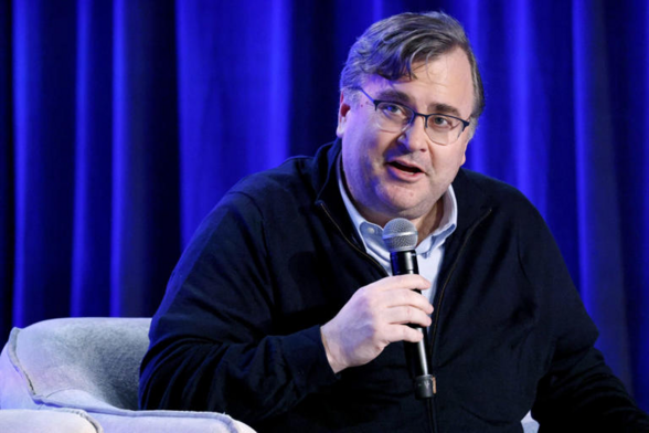 A picture of Reid Hoffman, with a blue draped backdrop, sitting on a chair. Wide-faced, overweight, in a button up and glasses. Major swoop of short hair. Middle aged.