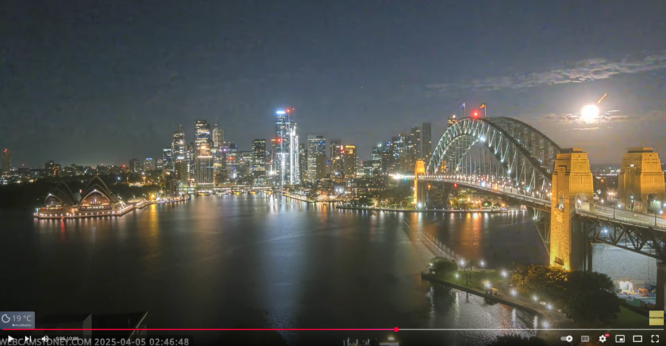A view of Sydney harbour and the city of Sydney at night time. The buildings and the harbour bridge are illuminated with city lights. On the right, a large fireball is occurring in the sky as the meteor plunges through the atmosphere.