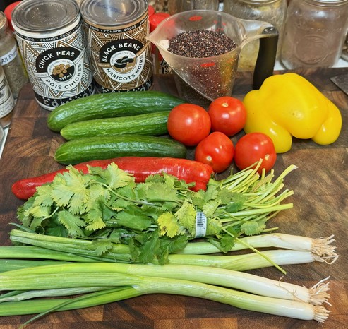 A cutting board on which are seen a can of chickpeas, a can of black beans, a clear plastic measuring cup holding black quinoa, 3 Persian cucumbers, 4 small tomatoes, 1/2 a large yellow bell pepper, a red sweet-tooth pepper, a small bunch of cilantro, and 4 thick scallions.