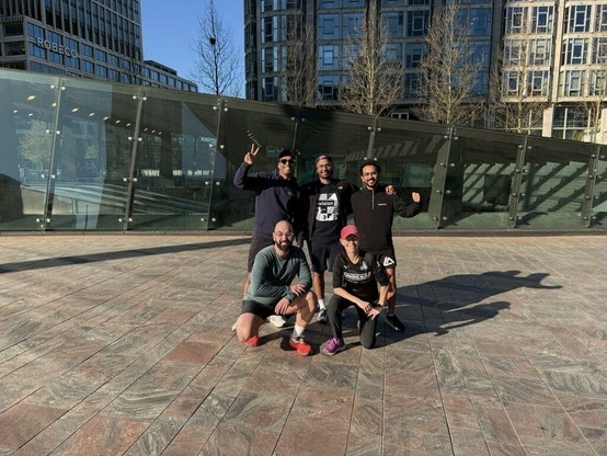Envision team members - Karthik - Bhagya - Ferkan - Amogh and Vanessa posing outdoors in a modern urban location outside Rotterdam Central Station. Three of the group are standing at the back and two kneeling in front. They are dressed in athletic wear, smiling and making celebratory gestures, as they have just completed a training run ahead of the Rotterdam Marathon. Behind them is a glass and metal structure with office buildings and leafless trees in the background, under clear sunny weather.