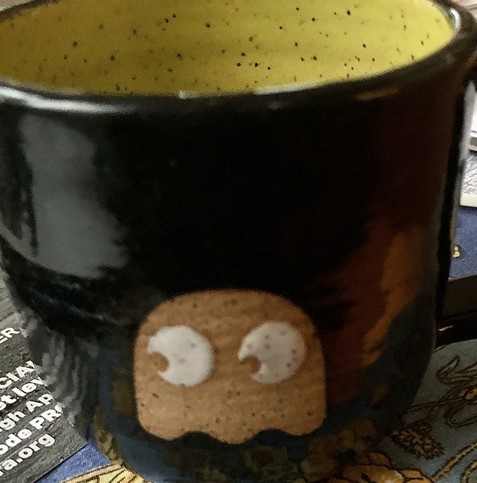 A dark blue (nearly black) mug with a yellow Ms. Pac-Man ghost on it and a yellow speckled interior, above some mail and a tablecloth.