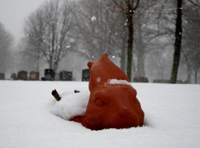 Photo of an orange/brown pottery bunny head poking up from beneath a soft blanket of newly fallen snow. Ears are held back in a typical signal of heightened awareness.  In the background are fresh snowflakes, tombstones, older leafless deciduous trees. A cemetery in Goderich, Ontario, (Huron Tract, Treaty 29, 1827 Ancestral land of Saugeen and Chippewas of Kettle and Stony Point First Nations.