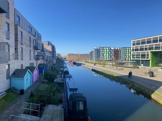 Two lovers stroll along the Union Canal amidst colorful beach huts, house boats, and colorful modern architecture