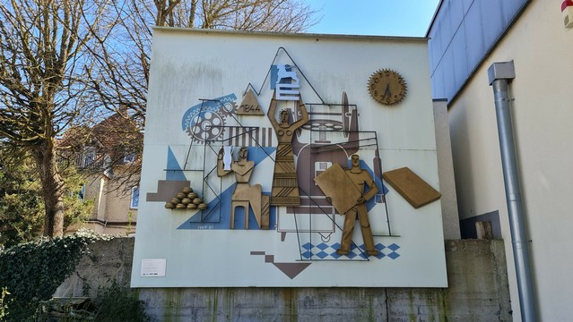 An Fassade angebrachte Reliefs aus blech und Drähten mit malerei dazu, stellt Maschinen, Arbeiter und gefertigte Produkte dar, Kannen, Räder, zeugs.