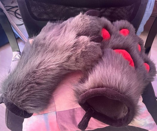 A photo of two sets of freshly sewn handpaws. They are made with gray fur, have black cuffs, and the pawpads are (a bit too...) red. They have four digits, and black claws made of minky fabric. The two paws are resting on a pillow, on a chair.