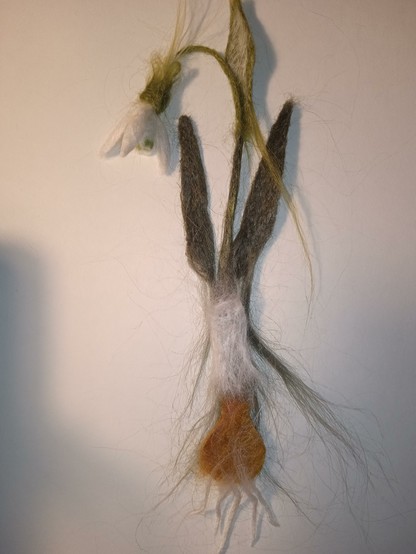 On a white background lies a snowdrop bookmark made from nettle using needle felting. It is not yet finished, the parts of the bookmark are superimposed on each other so that you can understand how it will look when it is finished. Thank you for your attention!