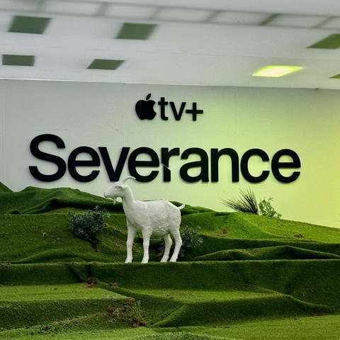 A white goat statue in on a stage of rolling green turf with stark white background featuring "Severance" and "Apple TV+" signs.