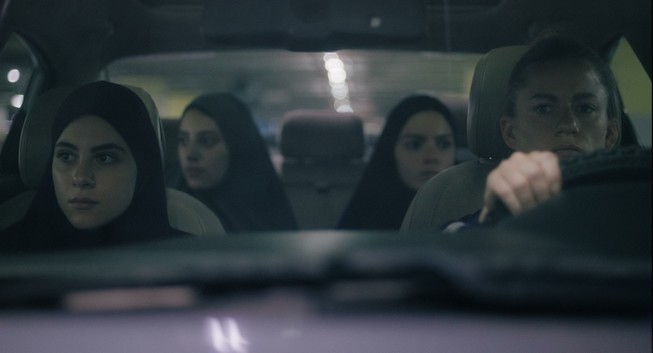 Die Innenansicht eines Autos. Am Steuer eine Frau mit blonden, zum Pferdeschwanz gebundenen Haaren, als Beifahrerinnen drei junge Frauen mit Hijab. Die Stimmung wirkt angespannt.