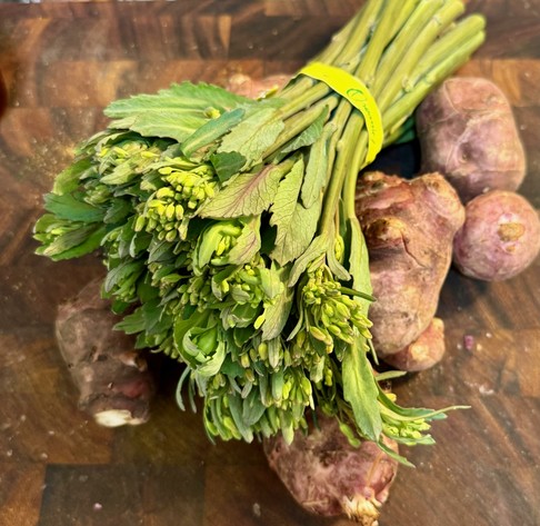 A bundle of leafy buds with a yellow band around the stems lying on reddish tubers on a cutting board.