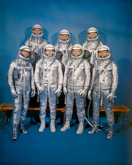Gruppenbild der "Mercury Seven" in silbrigen Raumanzügen. Die drei hinteren stehen auf einer einfachen Bierbank.