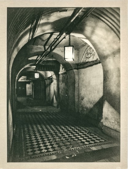 This image depicts a dimly lit, arched underground corridor of an old building. The corridor has curved ceilings with exposed pipes running along its length, and the walls appear aged and worn, showing signs of decay and grime. Two hanging square lanterns cast dramatic shadows, enhancing the mysterious, almost eerie atmosphere of the space. The floor features a striking black-and-white checkerboard tile pattern, partially damaged and worn with time.

The image is a *lith print*, a photographic technique known for its high contrast, gritty texture, and subtle tones, especially in the highlights. It was printed in a darkroom, giving the photograph its characteristic moody and artistic quality, with deep blacks, glowing whites, and a slightly grainy texture that adds to the nostalgic and haunting feel of the scene.