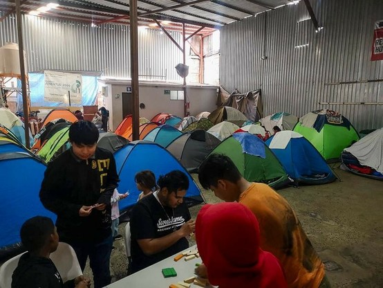 Das Foto zeigt einige Zelte, die in einer Wellblechhalle aufgebaut sind. Im Vordergrund sitzen Menschen an einem Tisch.