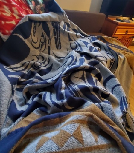 Close-up, medium shot of a blanket folded artfully over sofa. Predominant colors are blue, grey, and brown. The soft, diffused lighting emphasizes the fabric's texture and the detailed woven designs. The composition is slightly off-center, drawing attention to the mix of Coast Salish Indigenous patterns on the blanket. The background is blurred, creating shallow depth of field.