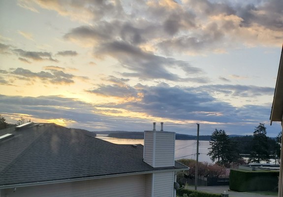 A tranquil scenic view of a sunrise over calm ocean waters. Houses form the foreground, with rooftops and chimneys visible. The landscape transitions into a calm expanse of water, beyond which are forested islands. The sky is filled with clouds streaked with gold.