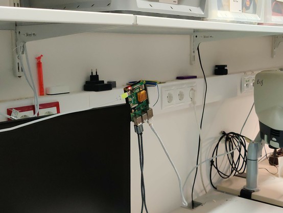 Ein eingeschalteter Raspberry Pi welcher wegen eines kurzen Ladekabels gefährlich an der oberen Ecke eines Bildschirms balanciert ist.