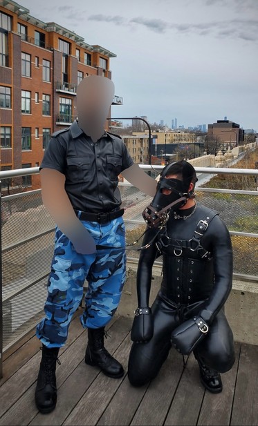 The same pony crouches down while kneeling on right knee. Sir stands to the pony’s right with Their right hand resting on the pony’s neck and Their left hand holding on the leather lead attached to a brown leather muzzle on the pony’s leather snout.
